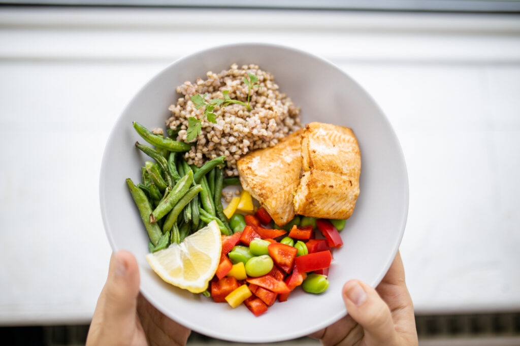 Hands holding salmon and buckwheat dish with green beans, broad beans, and tomato slices.