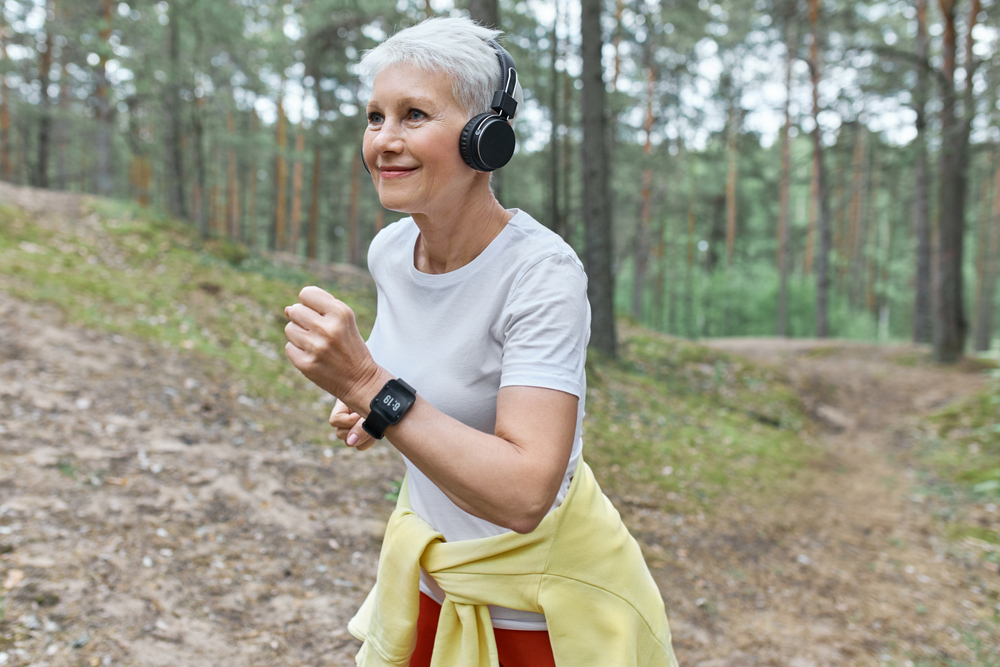 Older woman power walking in the woods with headphones on.