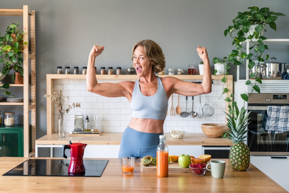 A fit older woman flexing arm muscles in the kitchen.