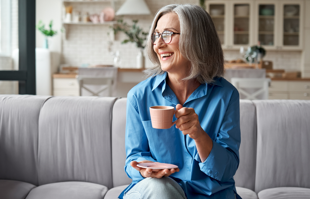 Happy older woman over 50 drinking coffee on the couch.