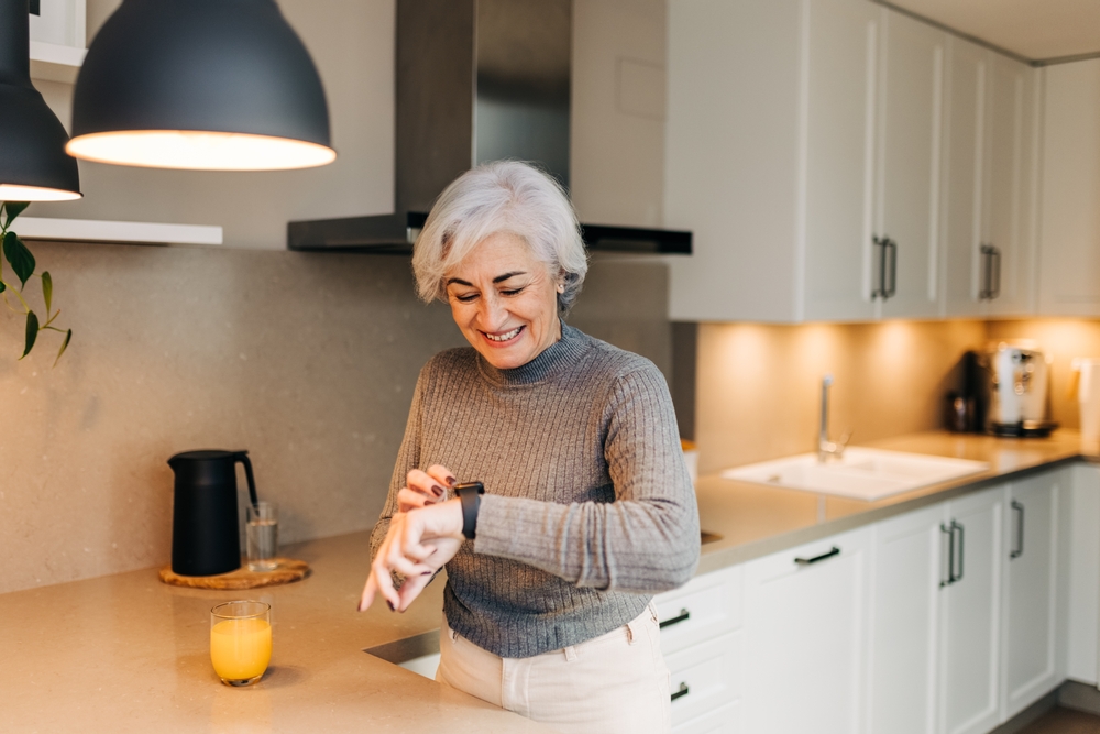 Healthy senior woman smiling at her smartwatch fitness tracker while standing in her kitchen.