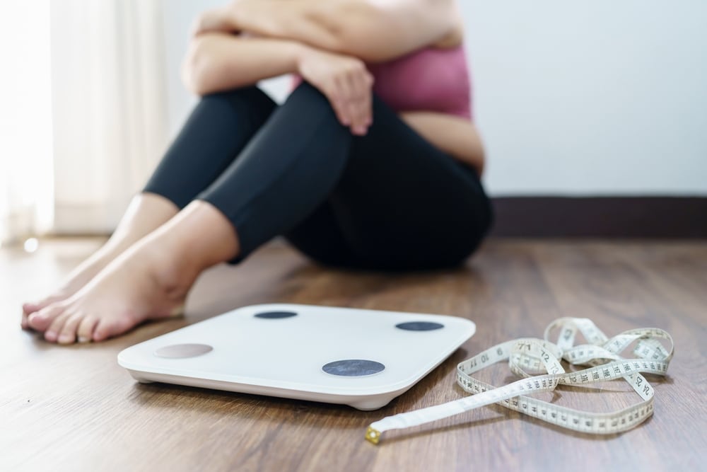 An overweight woman sitting on the floor next to a floor scale.