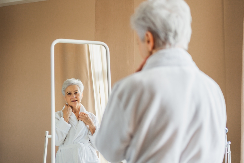 A senior woman in a bath robe looking at herself in the mirror.